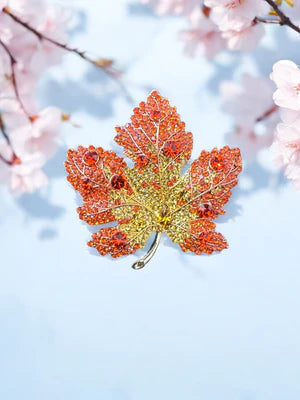 Decorative leaf brooch with red and gold accents against a cherry blossom background
