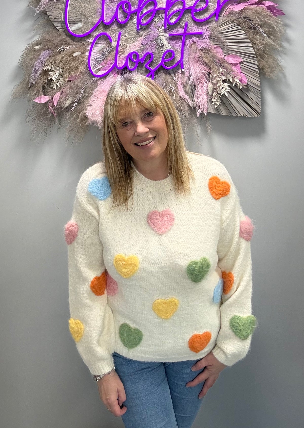 A woman wearing a cream-colored fuzzy knit sweater with colorful fuzzy heart patches in pink, orange, yellow, green, and blue, paired with blue jeans, standing in front of a gray wall with a decorative sign that reads 'Clover Closet'.