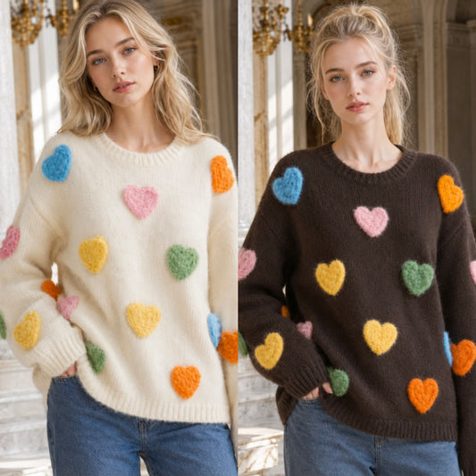 Two women modeling chunky knit sweaters with colorful fuzzy heart patches. The left sweater is cream with pastel blue, pink, yellow, green, and orange hearts.