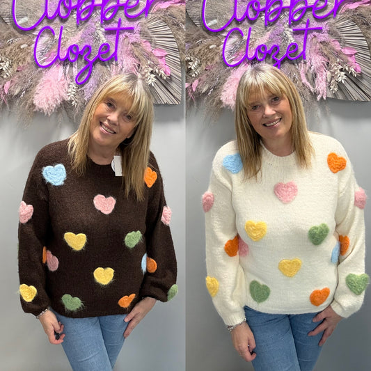 A side-by-side comparison of a woman modeling two fuzzy knit sweaters with colorful heart details. On the left, she wears a dark brown sweater with pastel blue, pink, yellow, green, and orange hearts.