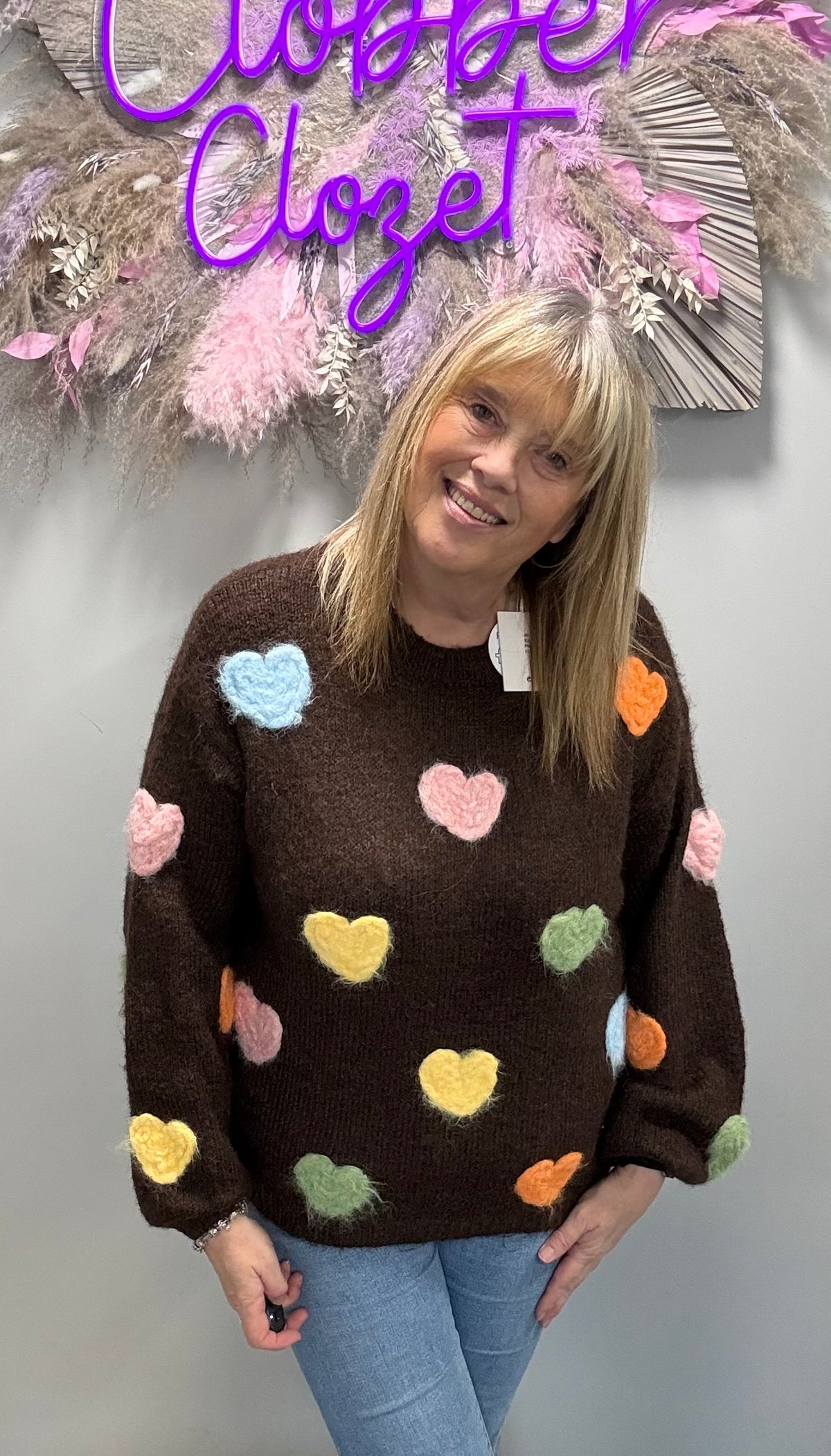 A woman wearing a brown fuzzy knit sweater with colorful pastel heart patches, paired with light blue jeans, standing in front of a gray wall with a decorative hanging and purple 'Closet Closet' text.
