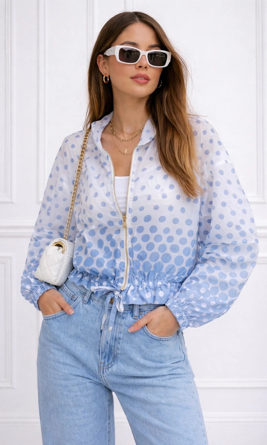 Woman wearing a light blue polka dot jacket and jeans with a white bag, against a white wall.