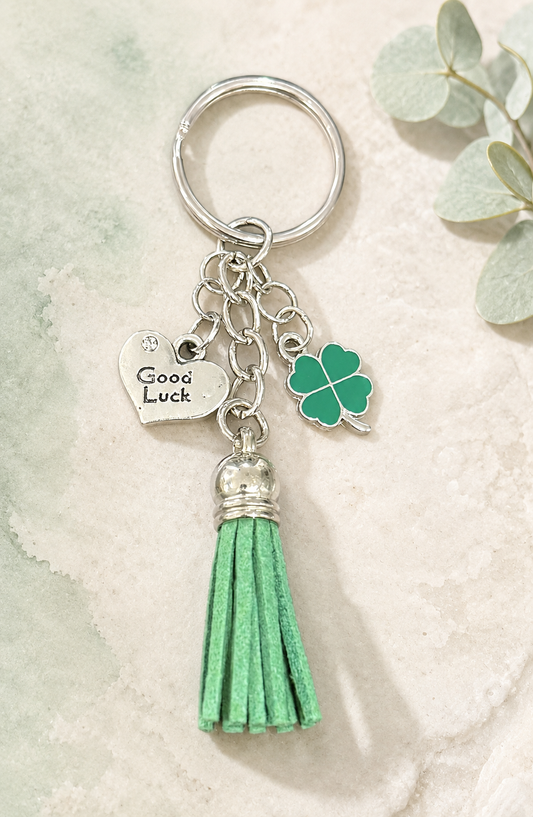 A silver-tone keychain with a split ring, featuring a heart charm engraved with 'Good Luck,' a green four-leaf clover charm, and a green tassel with a silver cap, displayed on a textured light background with decorative eucalyptus leaves.