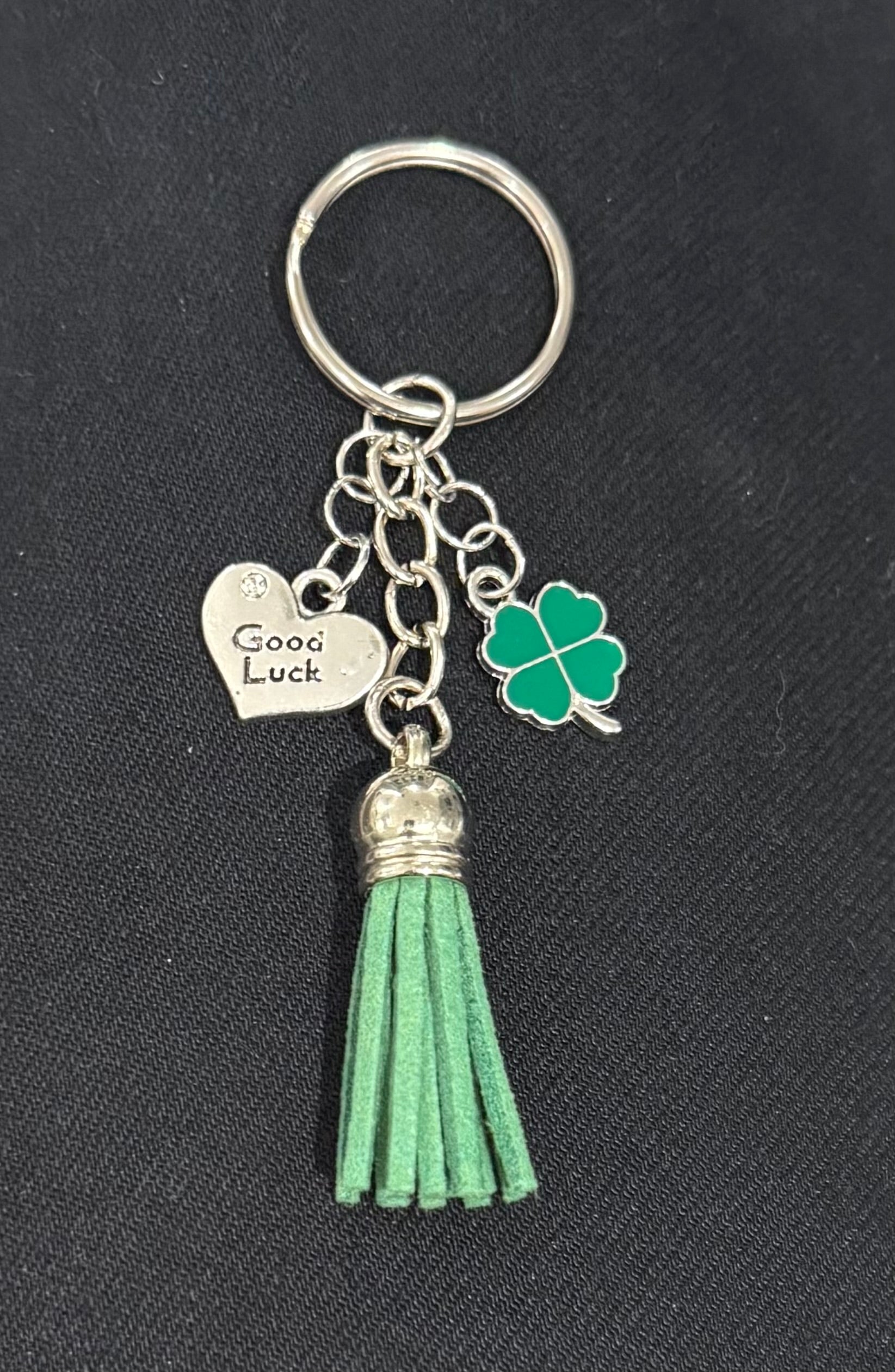 A St. Patrick's Day themed keychain featuring a green faux-leather tassel, a green four-leaf clover charm, and a silver heart charm engraved with 'Good Luck', all attached to a silver split ring on a black textured background.