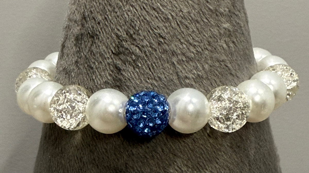 A beaded stretch bracelet displayed on a gray textured cone stand. The bracelet features white faux pearls, clear faceted crystal beads, and one central royal blue rhinestone-covered bead.