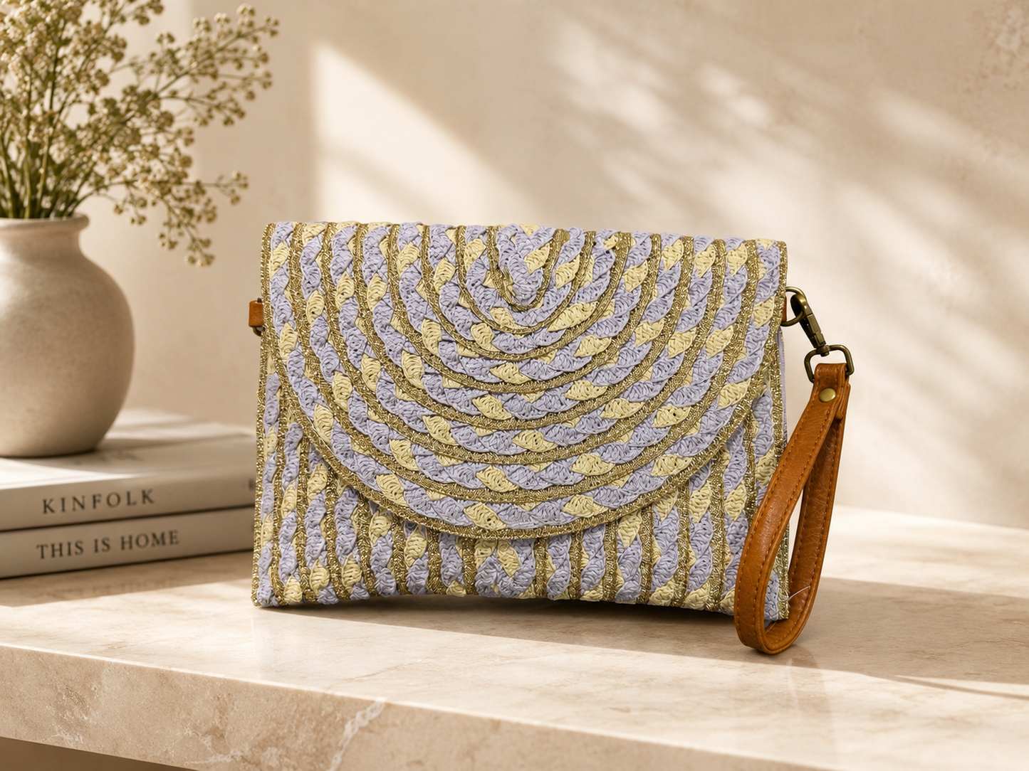 Patterned clutch bag with a brown strap on a light surface with books and a vase in the background
