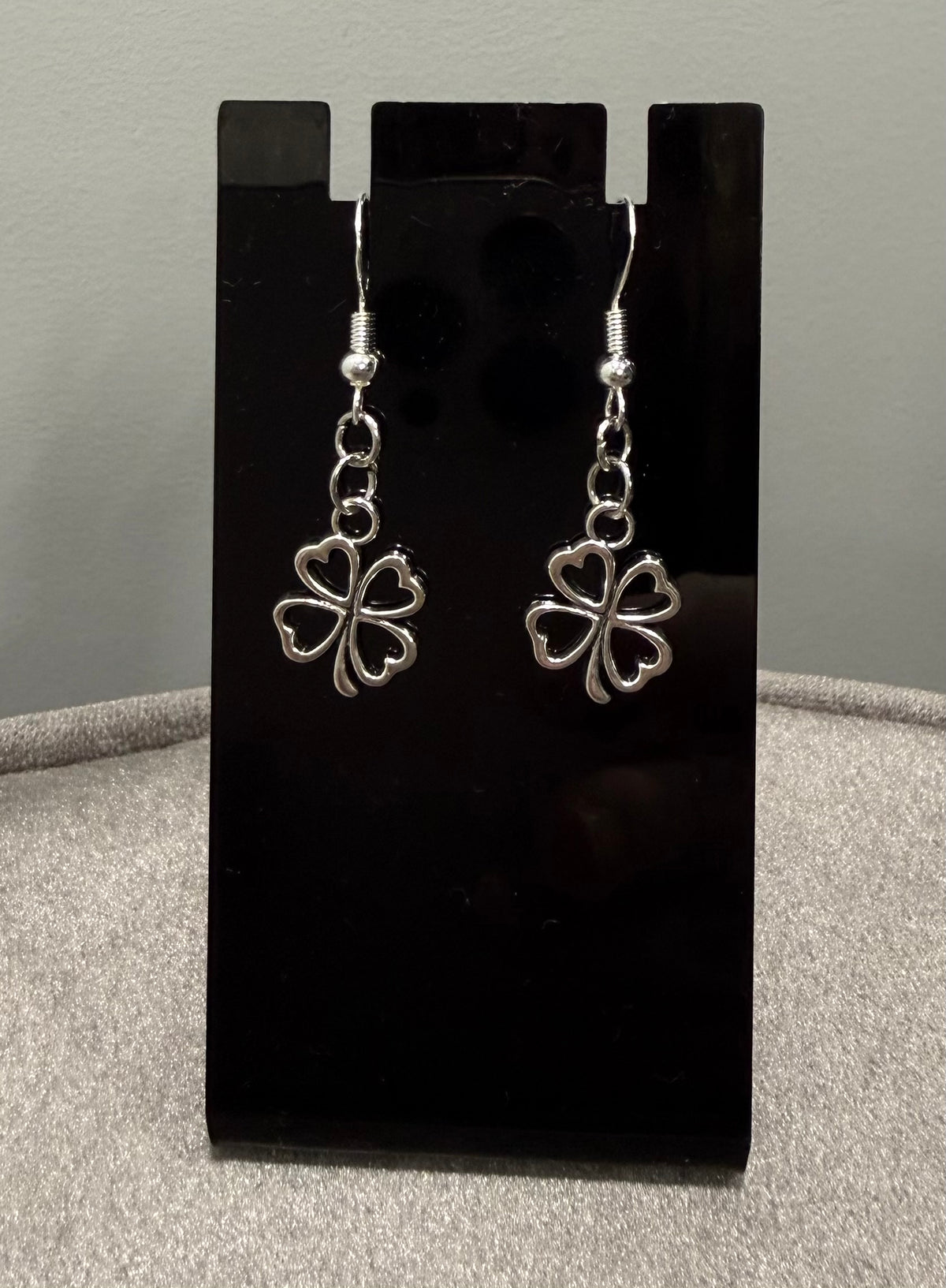 A pair of silver dangling earrings with four-leaf clover charms, displayed on a black stand against a gray background.