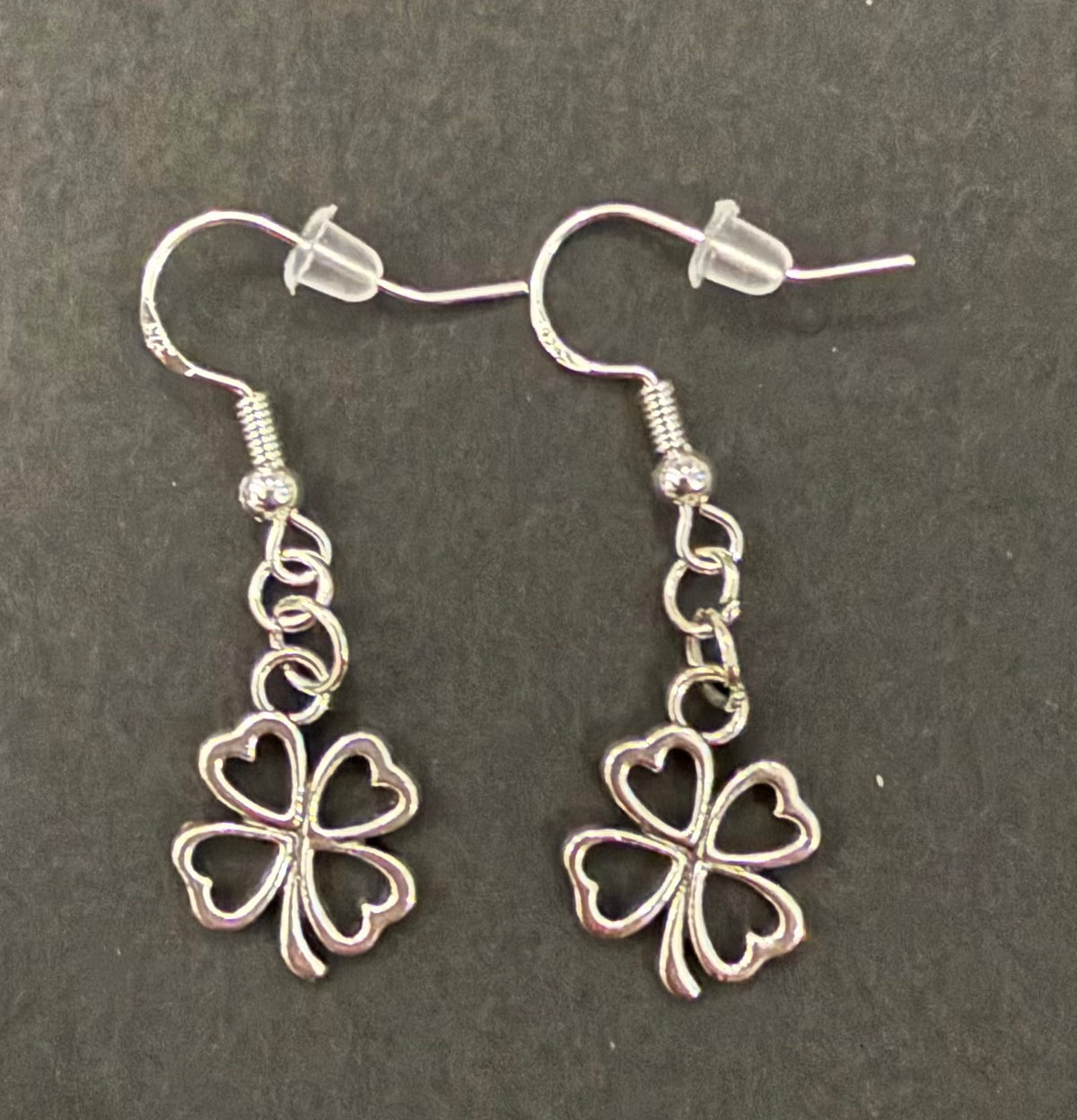 A pair of silver dangle earrings featuring openwork four-leaf clover charms with heart-shaped cutouts, displayed on a dark textured background.