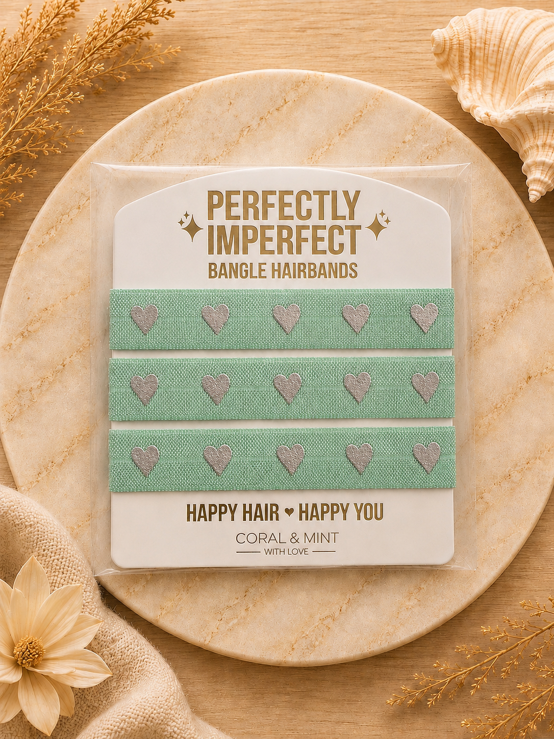 A packaged set of three mint green bangle hairbands with silver glitter hearts, displayed on a round beige stone plate with decorative dried flowers and a seashell.