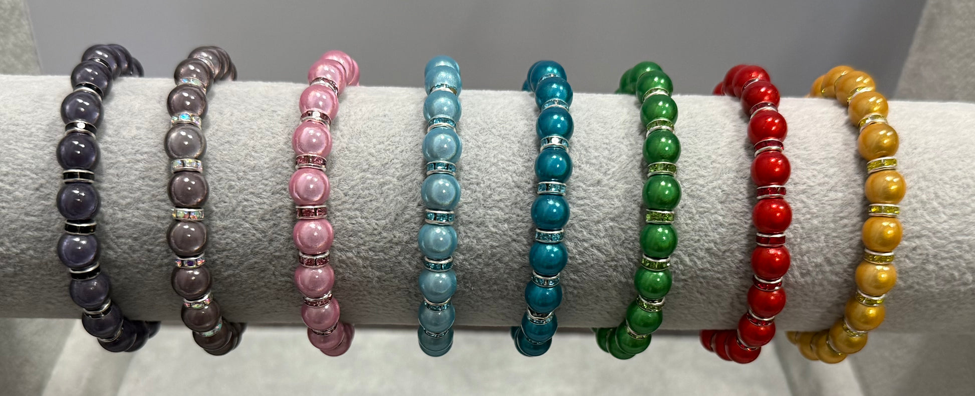 Eight beaded stretch bracelets displayed on a white velvet ring stand, featuring black, gray, pink, light blue, teal, green, red, and yellow beads with colour spacer rings.