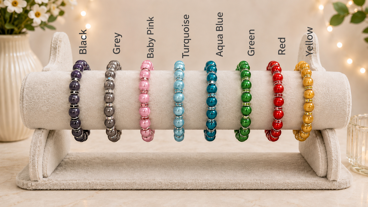 A display stand holding multiple beaded bracelets in different colors, labeled from left to right as Black, Grey, Baby Pink, Turquoise, Aqua Blue, Green, Red, and Yellow. The bracelets are arranged on a light beige velvet jewelry display against a softly blurred warm indoor background with string lights.