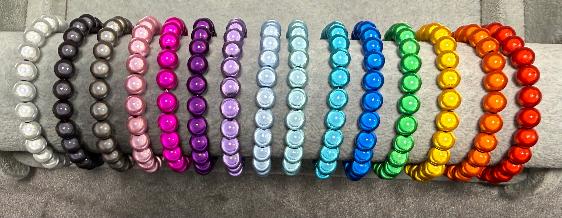A set of 16 beaded stretch bracelets arranged in two rows on a gray textured surface. The left row features metallic silver, dark gray, light pink, magenta, purple, and light blue bracelets.
