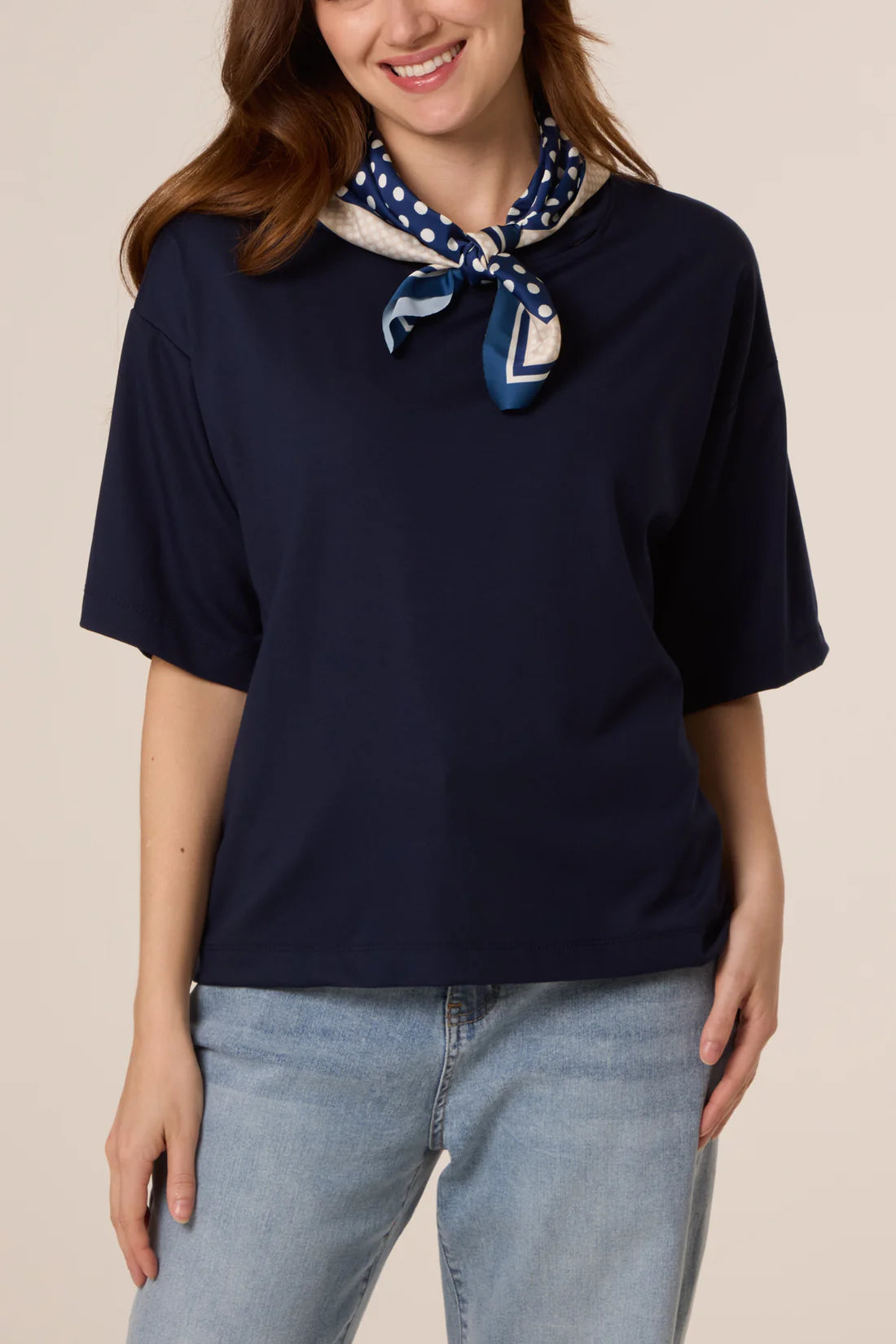 Woman wearing a navy blue top and light blue jeans with a patterned scarf around her neck on a beige background