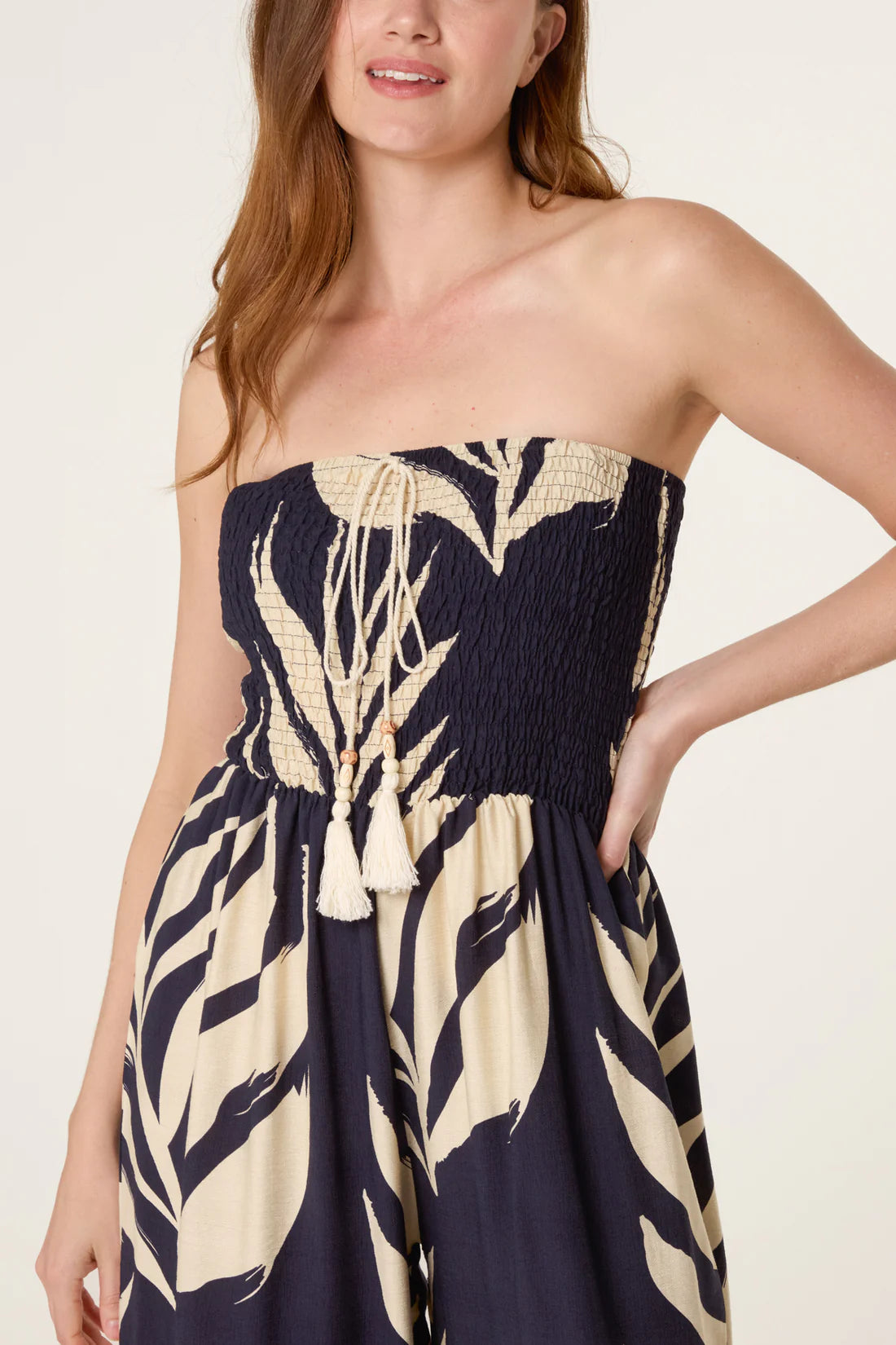 Woman wearing a strapless Navy Bandeau Tassel Leaf Print Jumpsuit with a leaf print pattern on a plain background