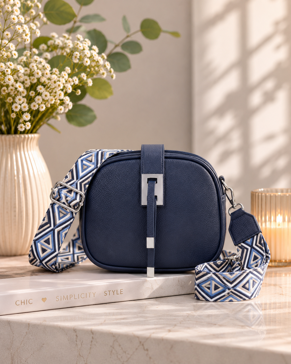 Navy blue handbag with a patterned strap on a marble surface with a vase of flowers and books in the background.