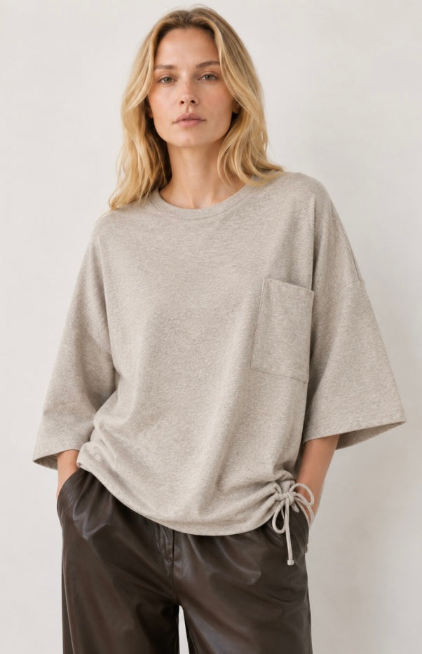 A woman wearing a light beige heathered knit top with wide three-quarter sleeves, a chest pocket, and a drawstring hem, paired with dark brown pants, against a plain light background.