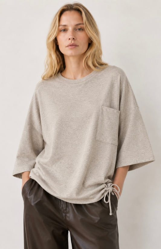 A woman wearing a light beige heathered knit top with wide three-quarter sleeves, a chest pocket, and a drawstring hem, paired with dark brown pants, against a plain light background.