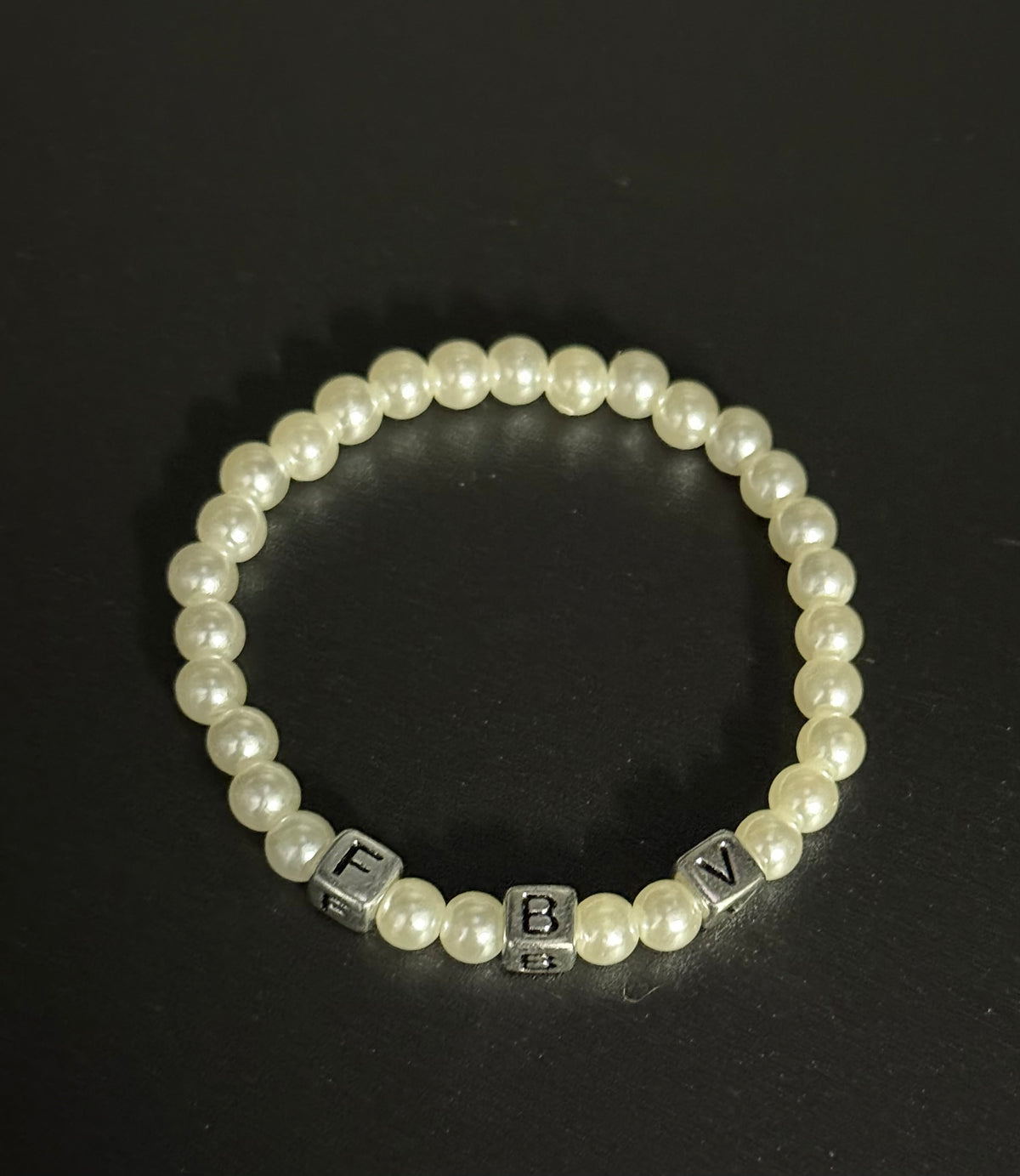 A beaded stretch bracelet featuring white faux pearls and silver cube letter initial beads F B and V, displayed on a dark black background.