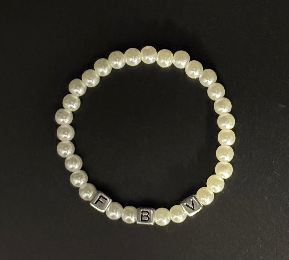 A beaded stretch bracelet featuring white faux pearls and silver letter beads spelling out 'F B V', displayed on a dark background.