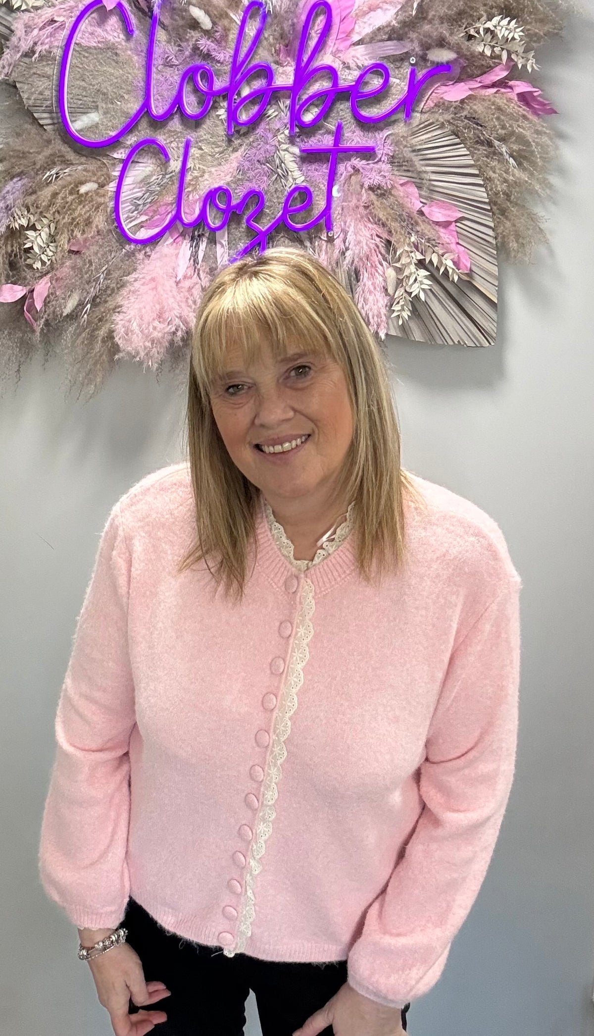 A woman wearing a light pink fuzzy cardigan with white lace trim and decorative buttons, standing in front of a gray wall with a decorative floral arrangement and a purple 'Closet' sign.