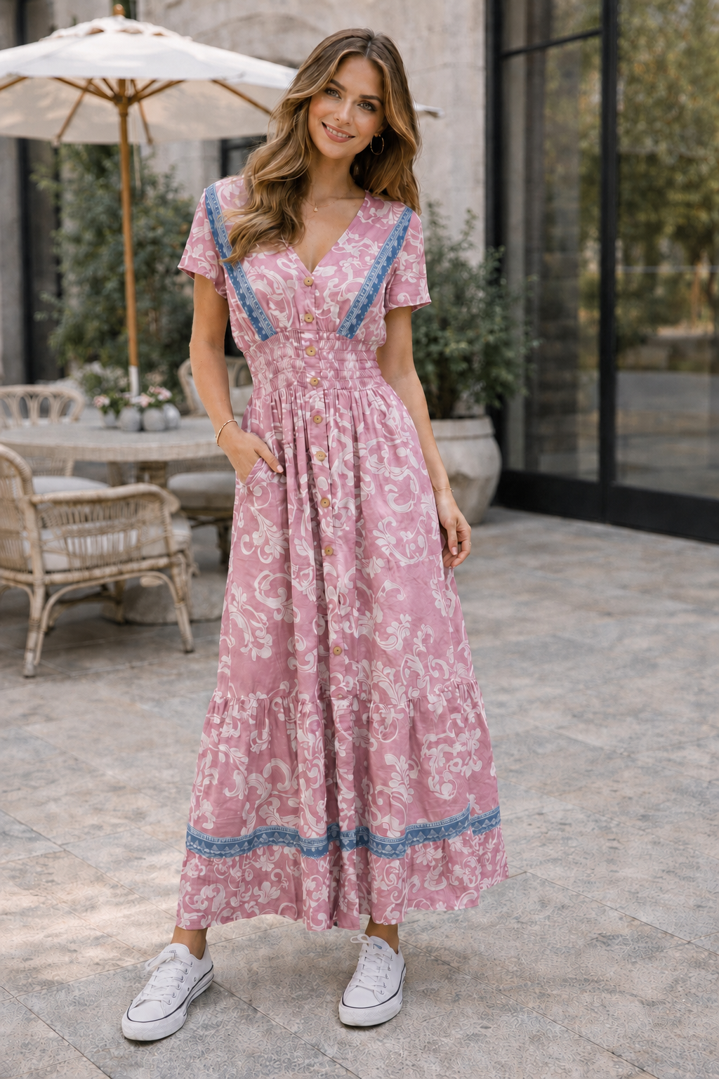 A woman wearing a pink and white floral maxi dress with blue trim, standing on a stone patio in front of a modern building with large glass windows.