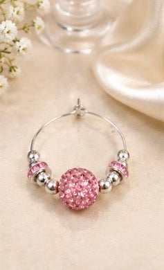 A silver wine glass charm adorned with a large pink glittery round bead and smaller pink and silver beads, displayed on a beige fabric surface with soft decorative props.