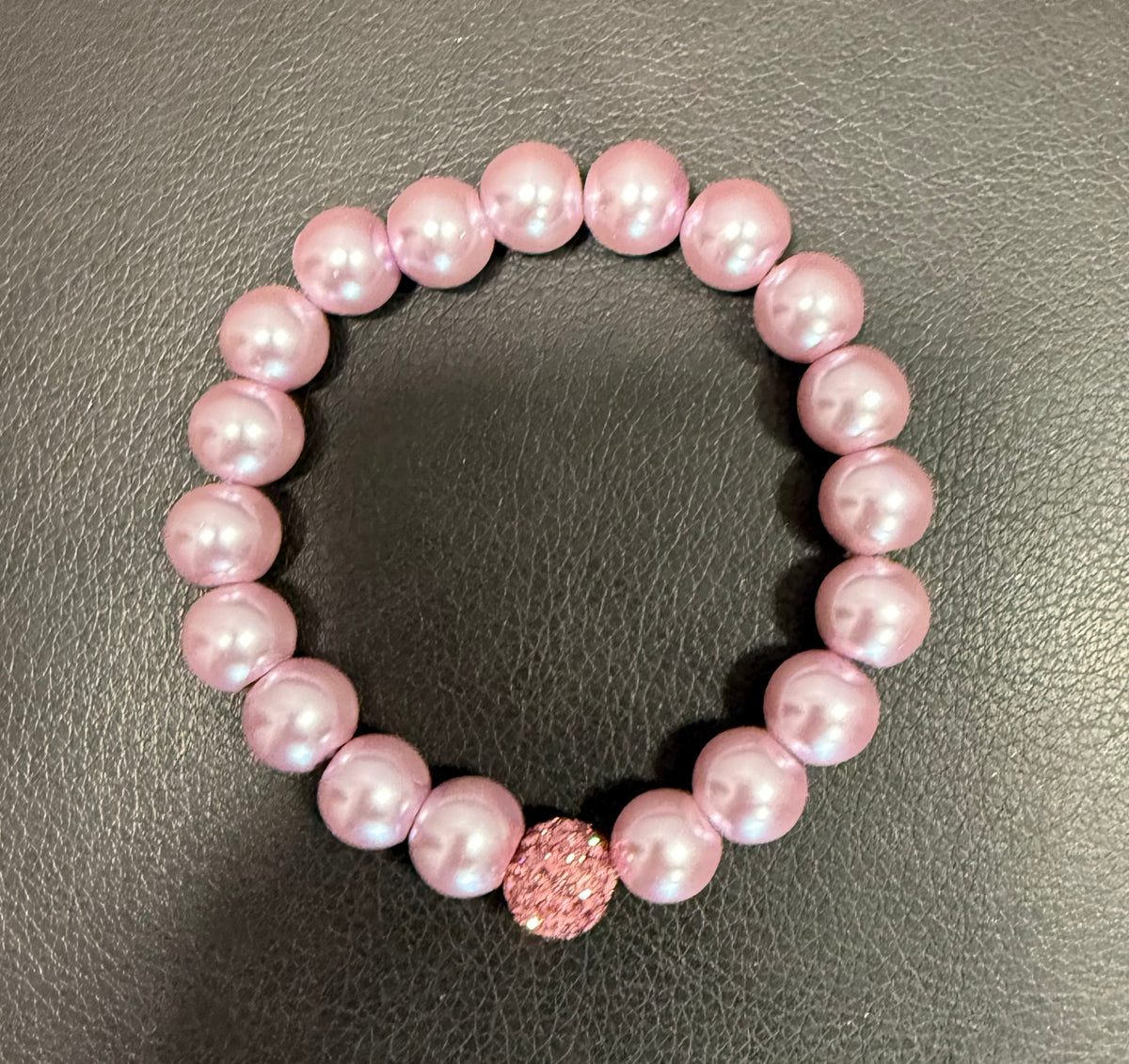 A pink beaded stretch bracelet featuring round lustrous pink pearls and a textured pink crystal accent bead, displayed on a dark gray textured leather background.