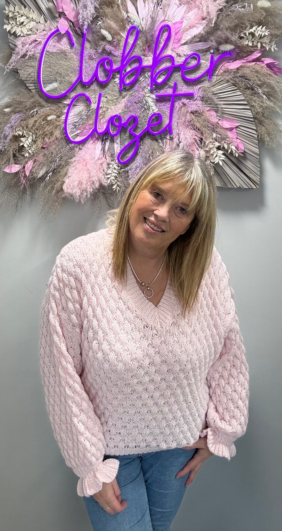A woman wearing a light pink textured knit sweater and blue jeans, standing in front of a gray wall with a decorative floral wreath and a purple 'Clobber Closet' sign.