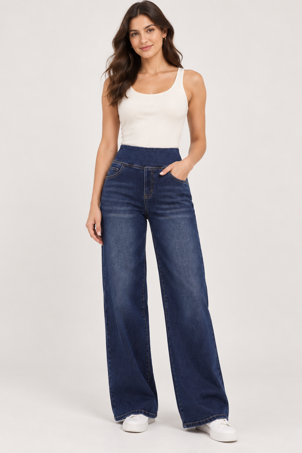 A woman modeling a pair of dark blue wide-leg jeans with a white ribbed tank top and white sneakers, standing against a light gray studio background.