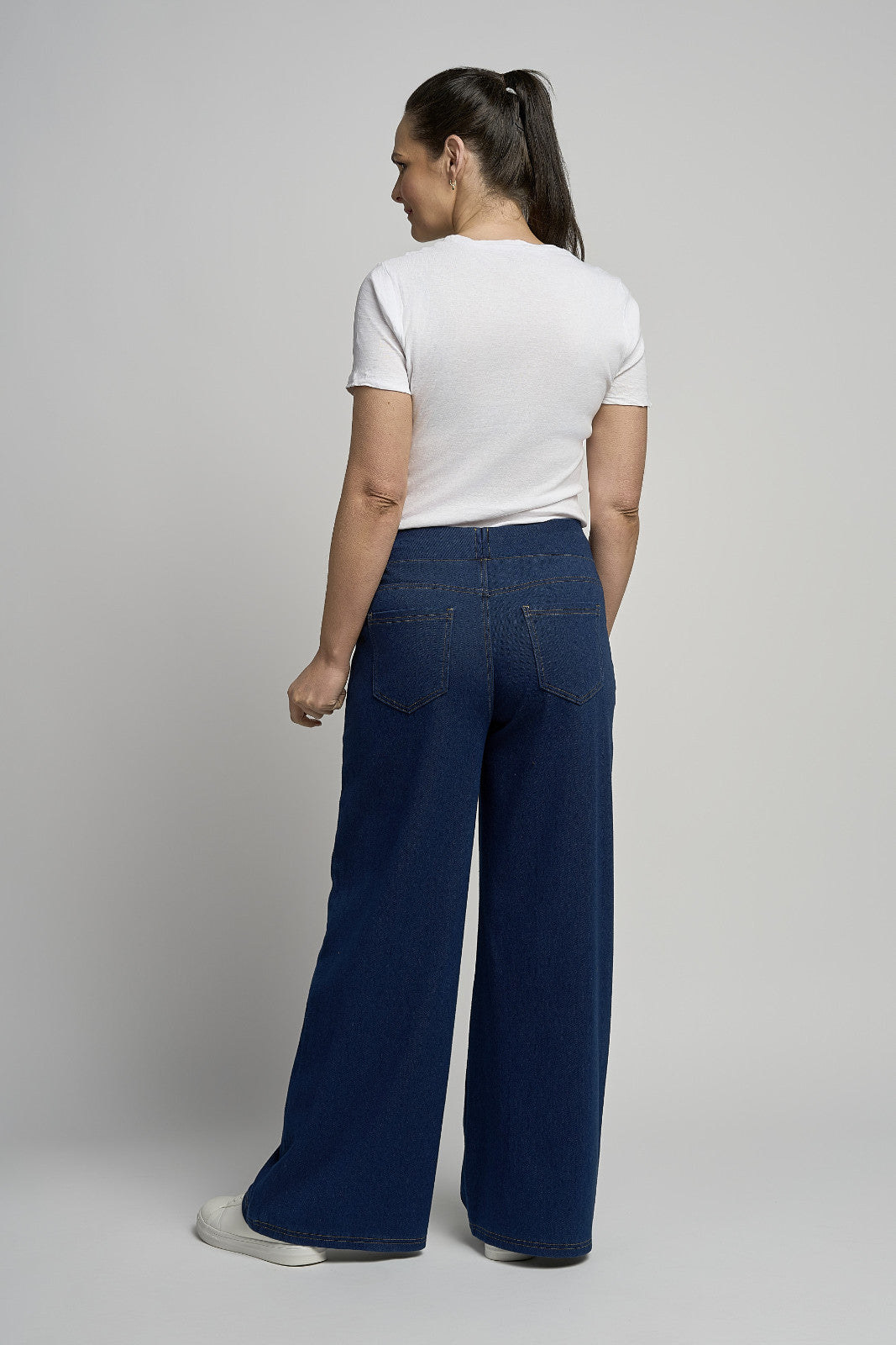 A woman shown from the back wearing a white short-sleeve t-shirt and dark blue wide-leg jeans, standing against a light gray studio background.