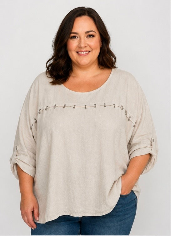 A woman modeling a light beige linen-blend top with rolled three-quarter sleeves and a decorative horizontal line of small metallic studs across the chest, paired with blue jeans against a light gray studio background.