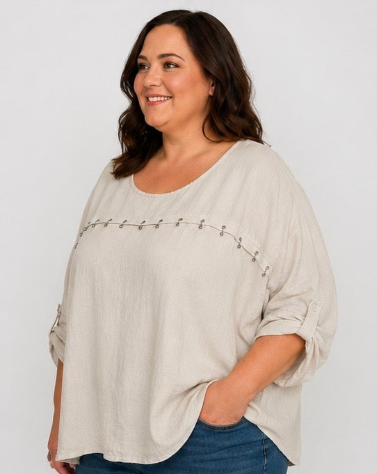 A woman wearing a light beige linen-blend top with rolled sleeves and a decorative line of silver studs across the chest, paired with blue jeans, against a plain light gray background.