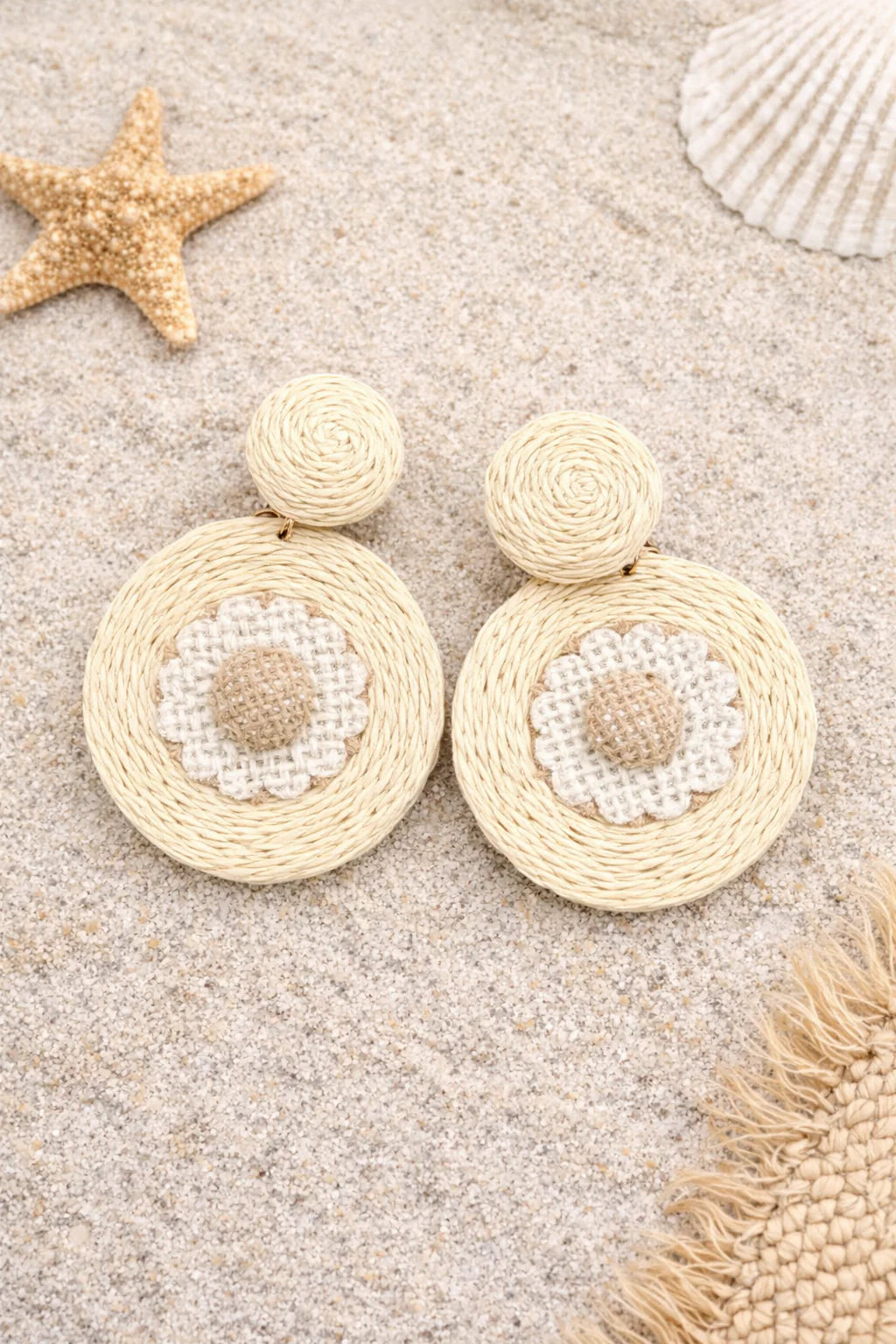 Beige raffia earrings with pearl-like centers on a sandy background with starfish and shells.