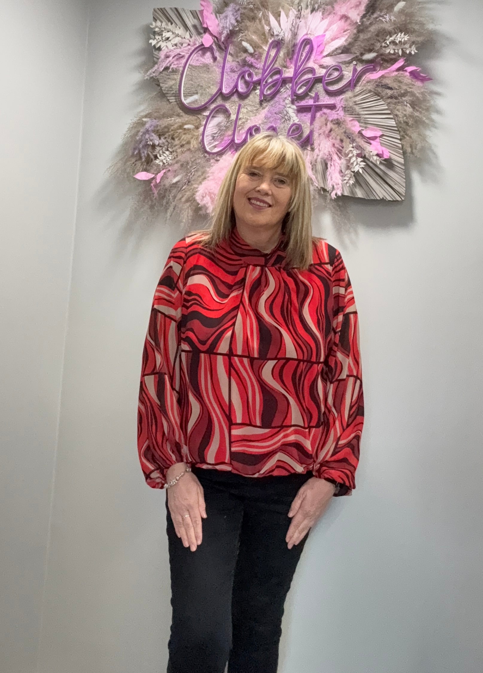 Person wearing a red and black patterned top standing in front of a decorative wall with 'Clobber Clozet' sign.