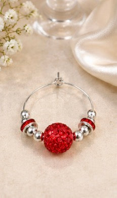 A silver-tone beaded wine glass charm with a large, round, sparkling red crystal bead at the center, displayed on a soft beige fabric background with small white flowers and a glass stem in the background.