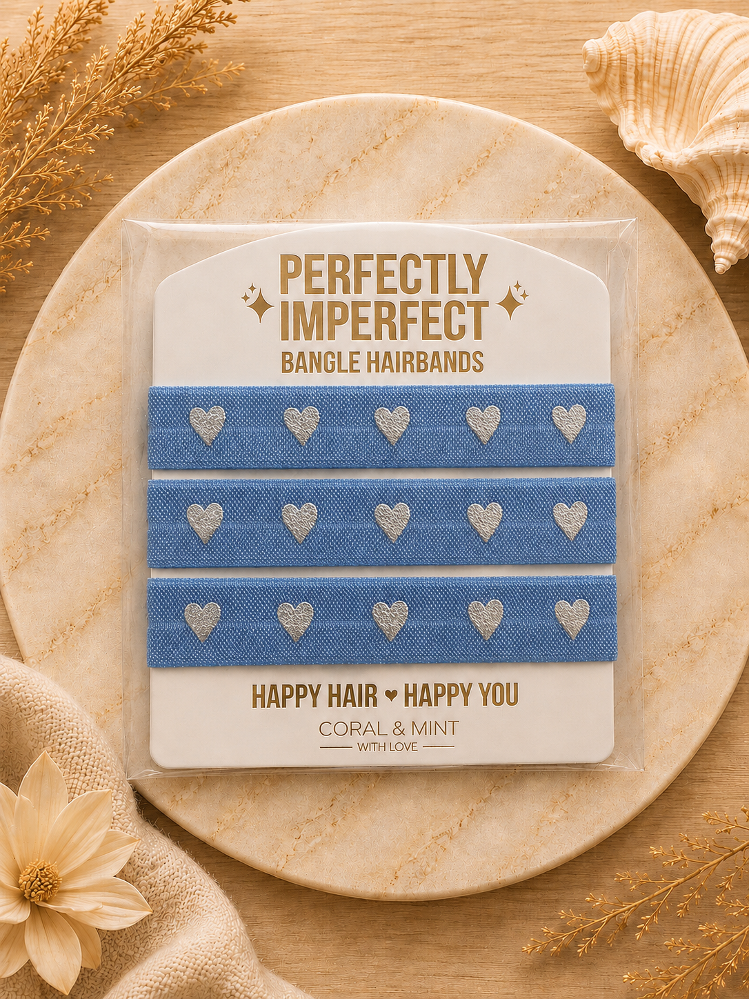 Three blue bangle hairbands with silver glitter hearts, packaged on a white card reading 'PERFECTLY IMPERFECT BANGLE HAIRBANDS' and 'HAPPY HAIR HAPPY YOU', displayed on a round stone tray with dried flowers and a seashell.