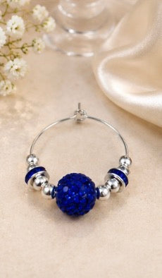 A silver-tone wine glass charm with a central round blue beaded cluster, displayed on a beige fabric surface with soft decorative flowers and satin in the background.