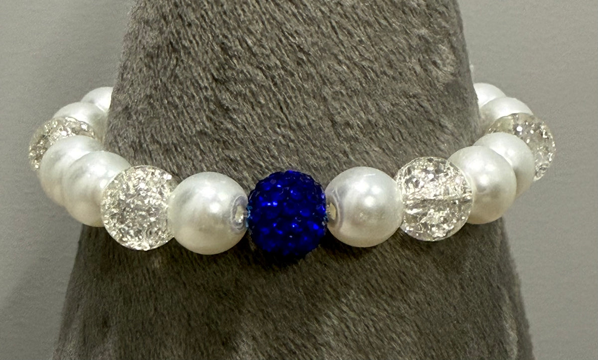A beaded stretch bracelet displayed on a gray textured stand, featuring white faux pearls, clear faceted beads, and a central royal blue rhinestone bead.