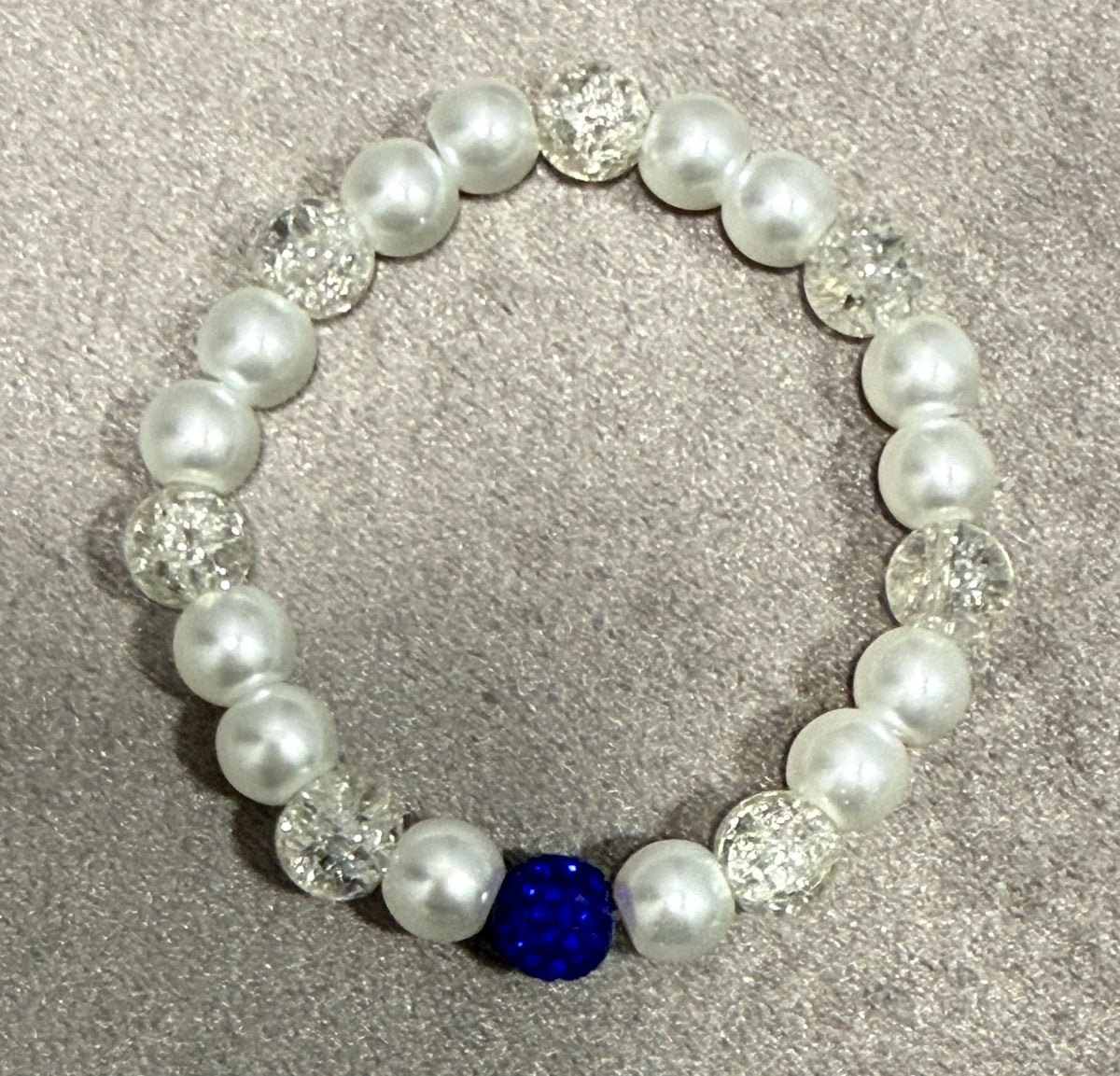 A beaded stretch bracelet featuring white faux pearls, clear faceted crystal beads, and one prominent royal blue faceted bead, displayed on a textured gray background.