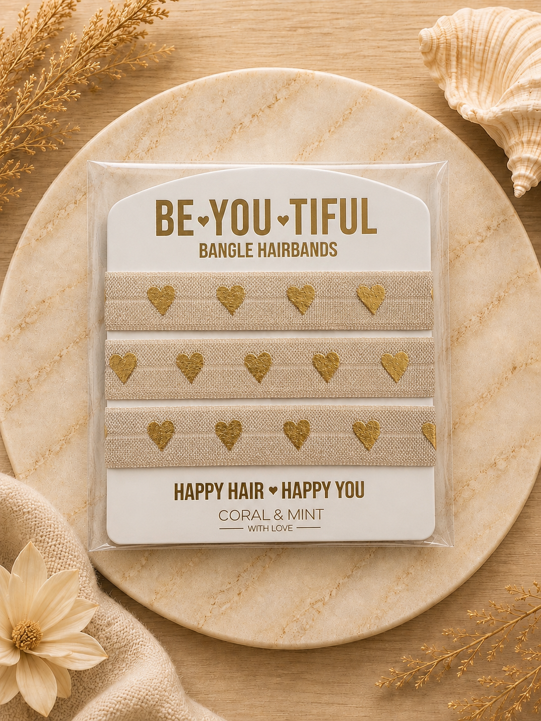A package of four bangle hairbands with gold heart patterns on a beige linen fabric, displayed on a round wooden tray with decorative dried flowers and a seashell.