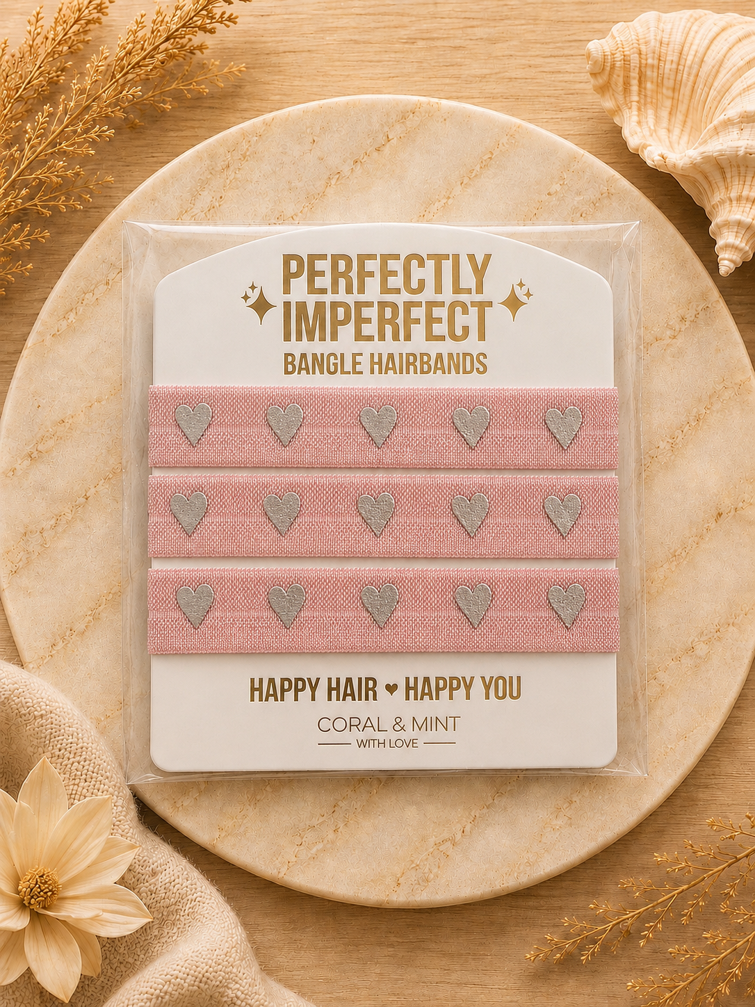 Pack of three pink bangle hairbands with silver glitter heart details, presented on a branded card. The card is placed on a round stone tray with decorative dried grass, a flower, and a seashell on a wooden surface.