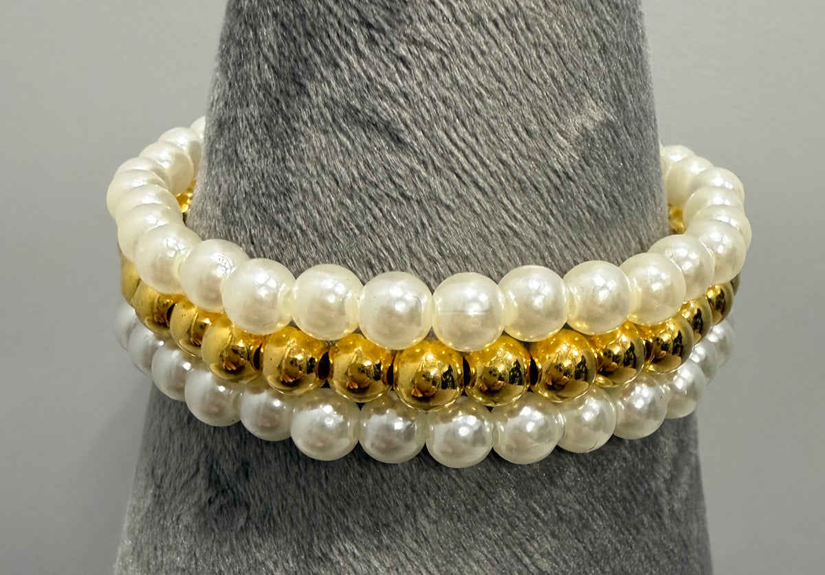 A stack of three beaded stretch bracelets displayed on a gray textured cone stand. The top bracelet is made of large white faux pearls, the middle bracelet features shiny gold metallic beads, and the bottom bracelet is a combination of white pearls and gold metallic beads.