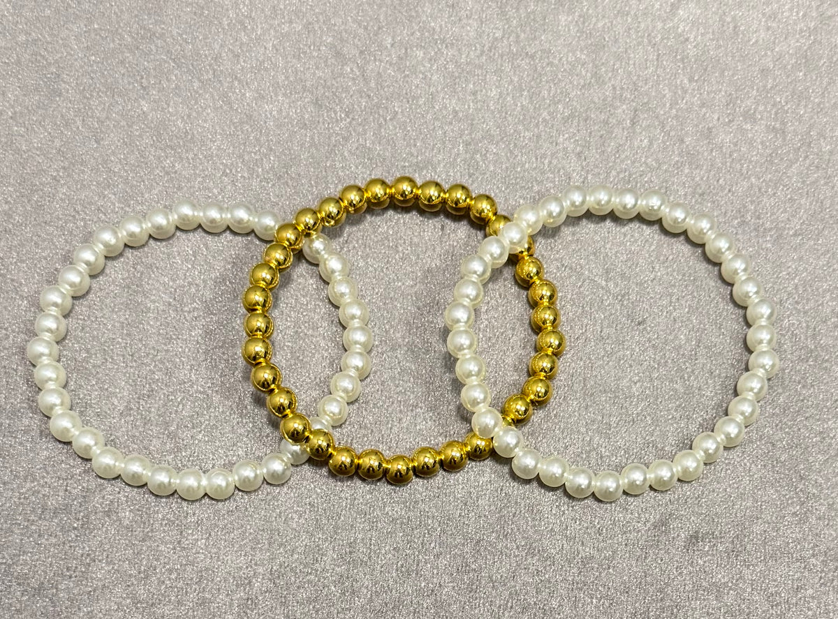 Three beaded stretch bracelets arranged in a circle on a textured gray background. Two bracelets are made of white faux pearls, and one is made of shiny gold beads.