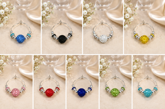 A collage of ten wine glass charms, each a silver-tone ring with a round faceted gemstone in a different color (blue, black, white/silver, yellow, pink, red, dark blue, green, light blue, and teal) and small metallic spacer beads, displayed on a beige satin fabric with white flowers and glassware in the background.