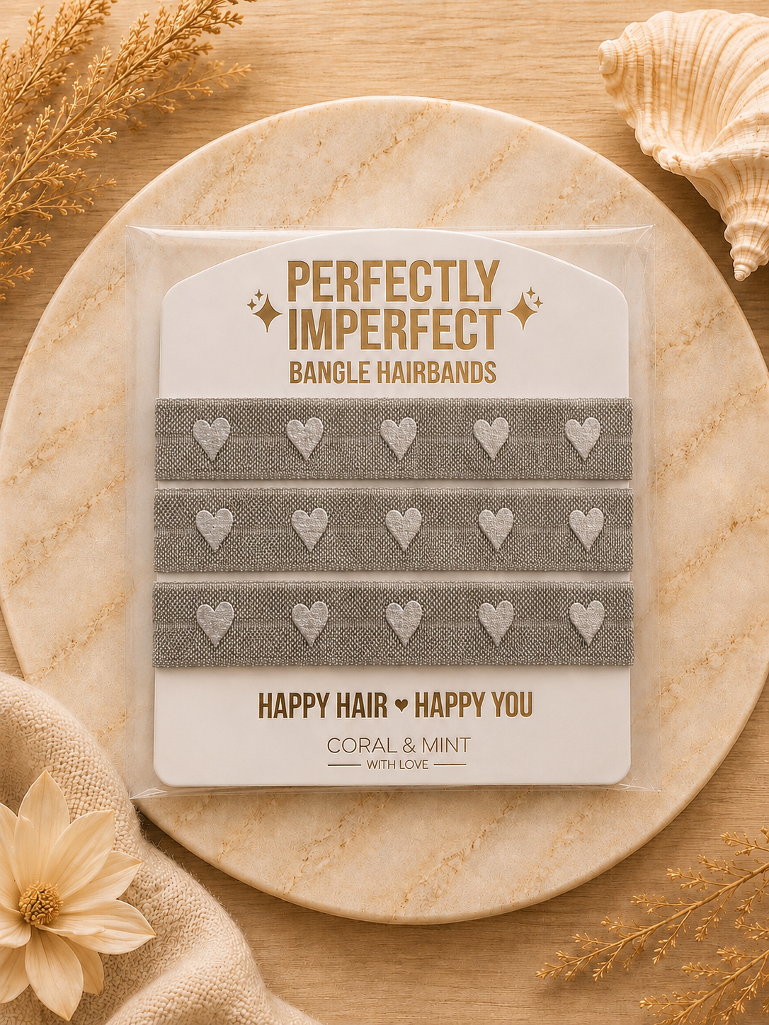 A package of four gray bangle hairbands with silver heart patterns, presented on a branded card. The package is styled on a round stone plate with dried flowers and a seashell on a wooden background.