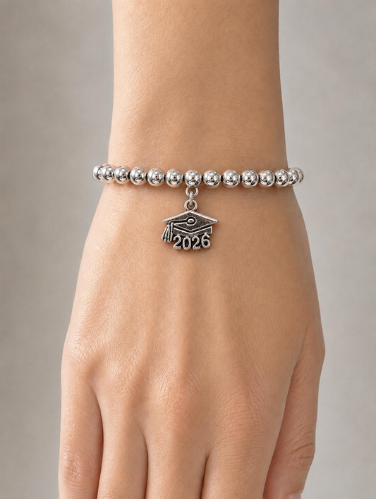 A silver beaded bracelet worn on a wrist, featuring a graduation cap charm with the year '2026' engraved on the front, shown against a neutral gray background.