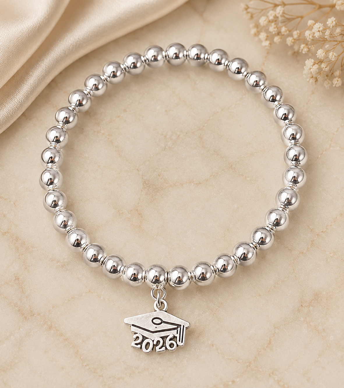 A silver beaded bracelet with a graduation cap charm engraved with '2026', displayed on a beige marble surface with a cream satin fabric and dried flowers in the background.