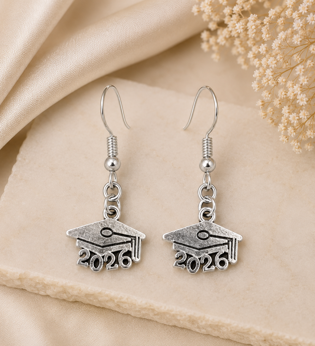 A pair of silver drop earrings shaped like graduation caps with the year 2026 engraved below, displayed on a beige fabric surface with dried flowers in the corner.