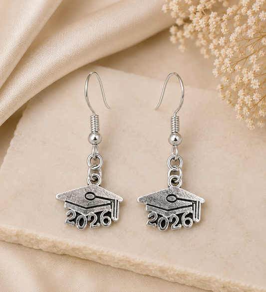 A pair of silver drop earrings shaped like graduation caps with the year 2026 engraved below, displayed on a beige fabric surface with dried flowers in the corner.