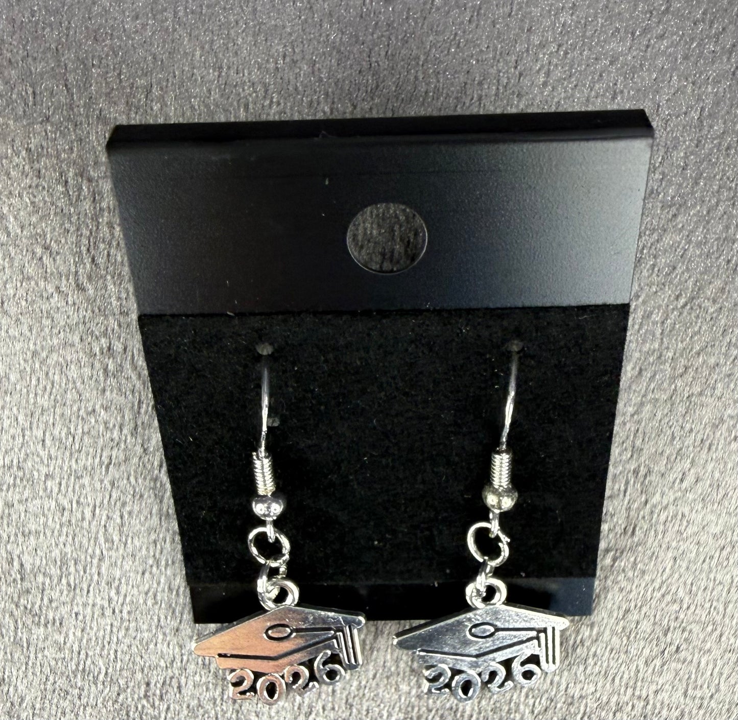 A pair of silver dangling earrings shaped like graduation caps with '2026' cut out at the bottom, displayed on a black card against a gray textured background.